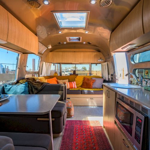 The image shows a cozy, modern RV interior with a seating area, kitchen, and dining space accented by colorful cushions.