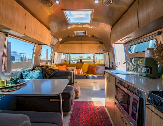 The image shows a cozy, modern RV interior with a seating area, kitchen, and dining space accented by colorful cushions.