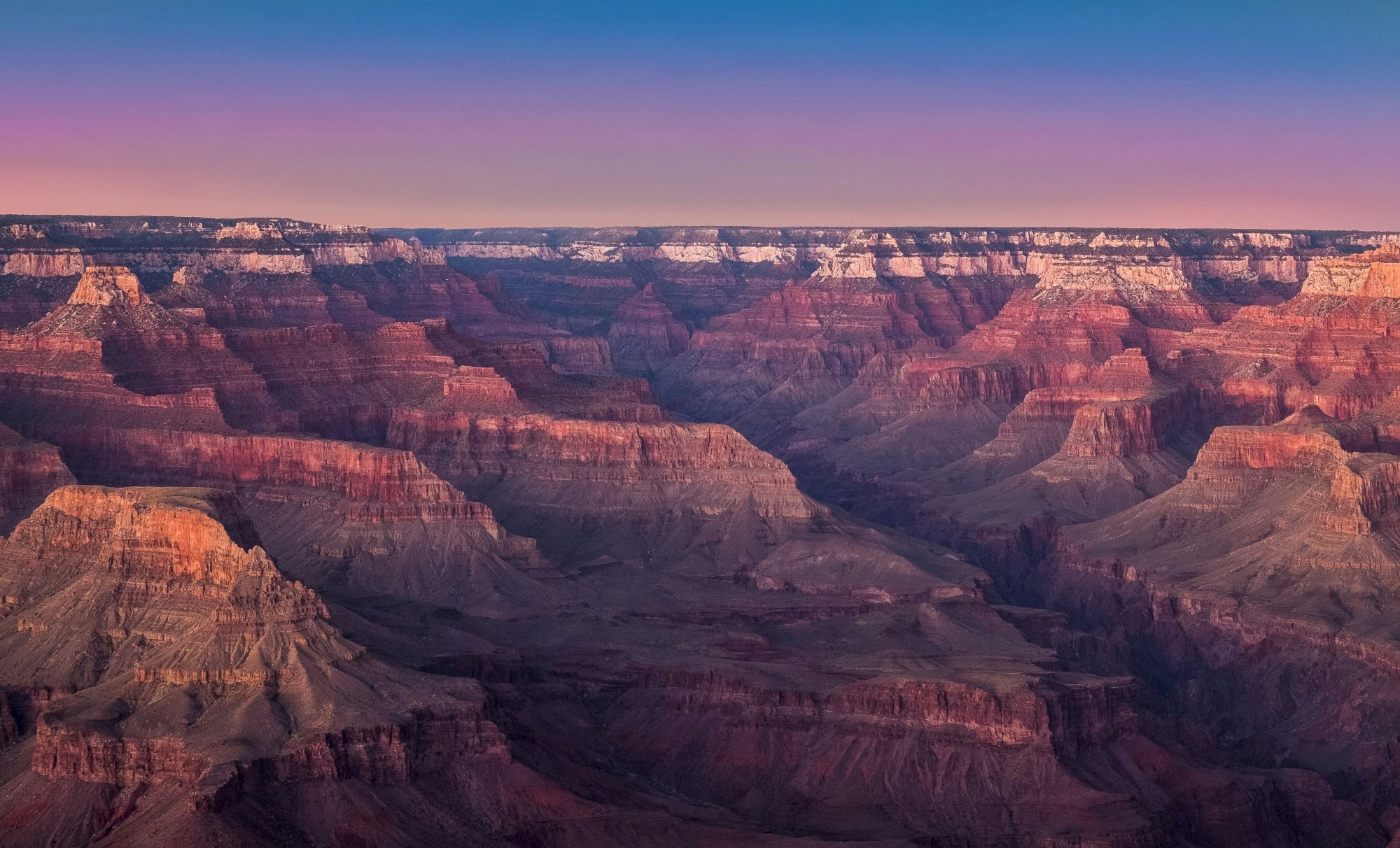A vast, colorful landscape of the Grand Canyon at sunset with deep valleys and layered rock formations under a gradient sky.