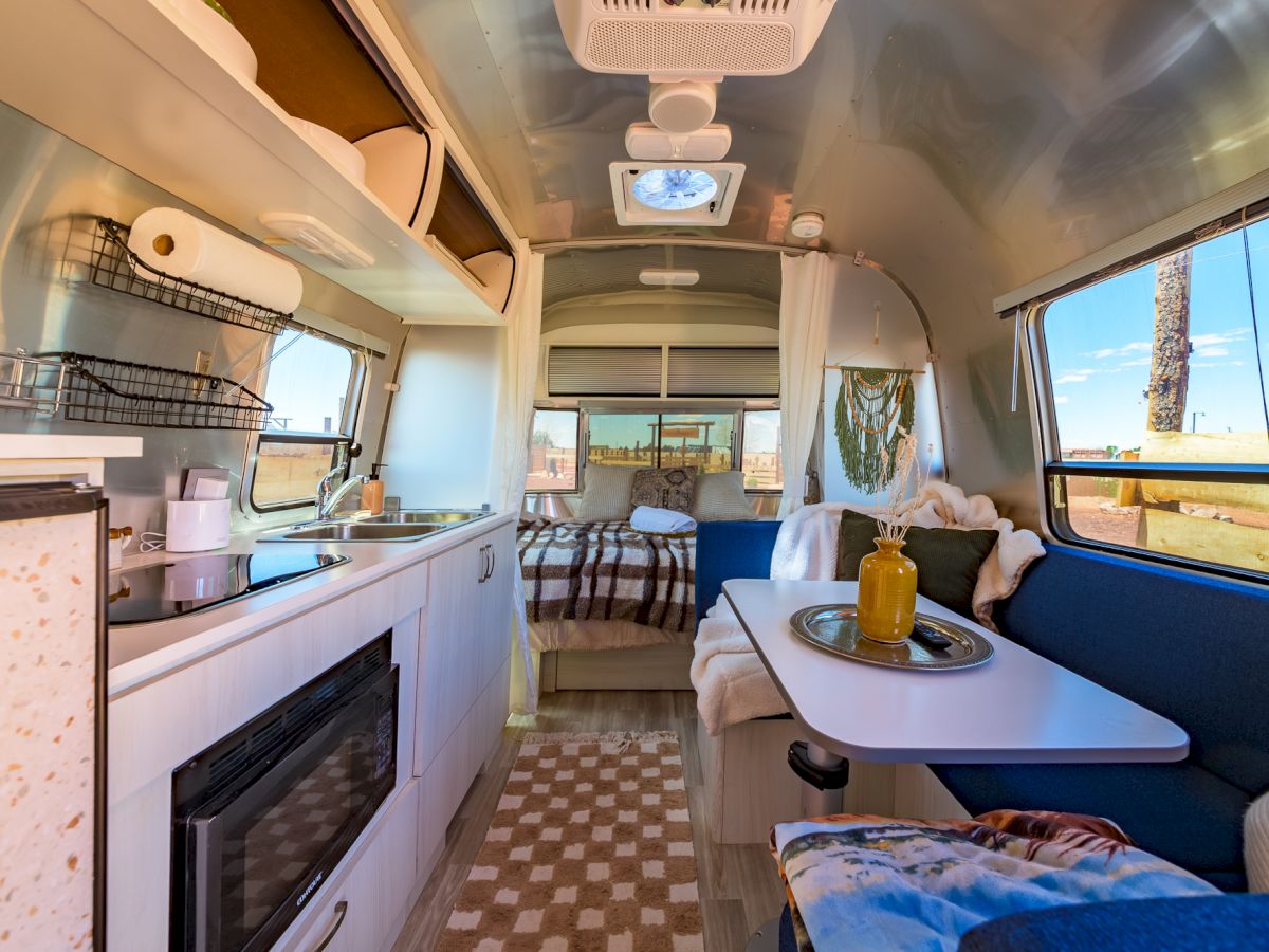 The image shows the interior of a modern RV with a kitchen, seating area, and bed, featuring cozy decor and a bright, spacious feel.