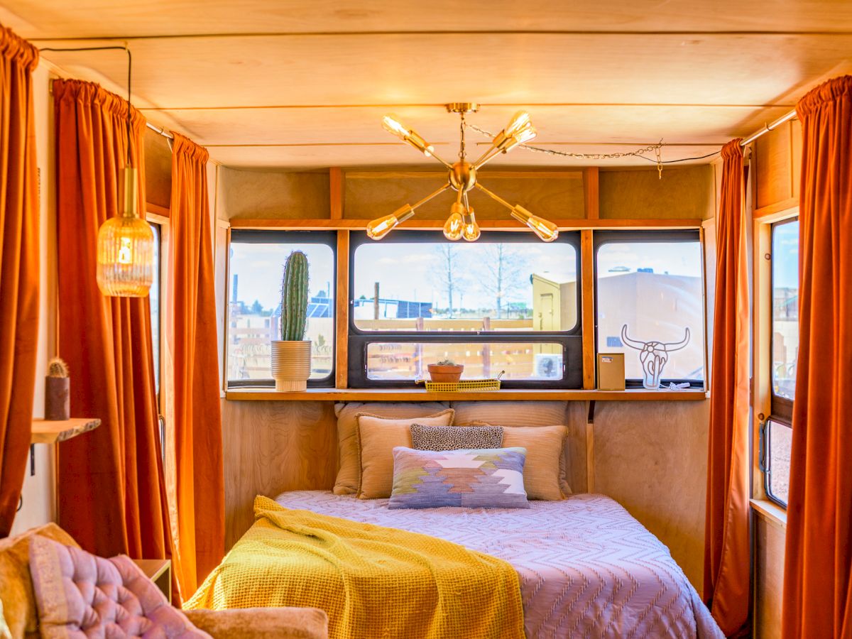 A cozy room with a bed, vibrant orange curtains, modern lighting, and large windows letting in natural light and a view outside.