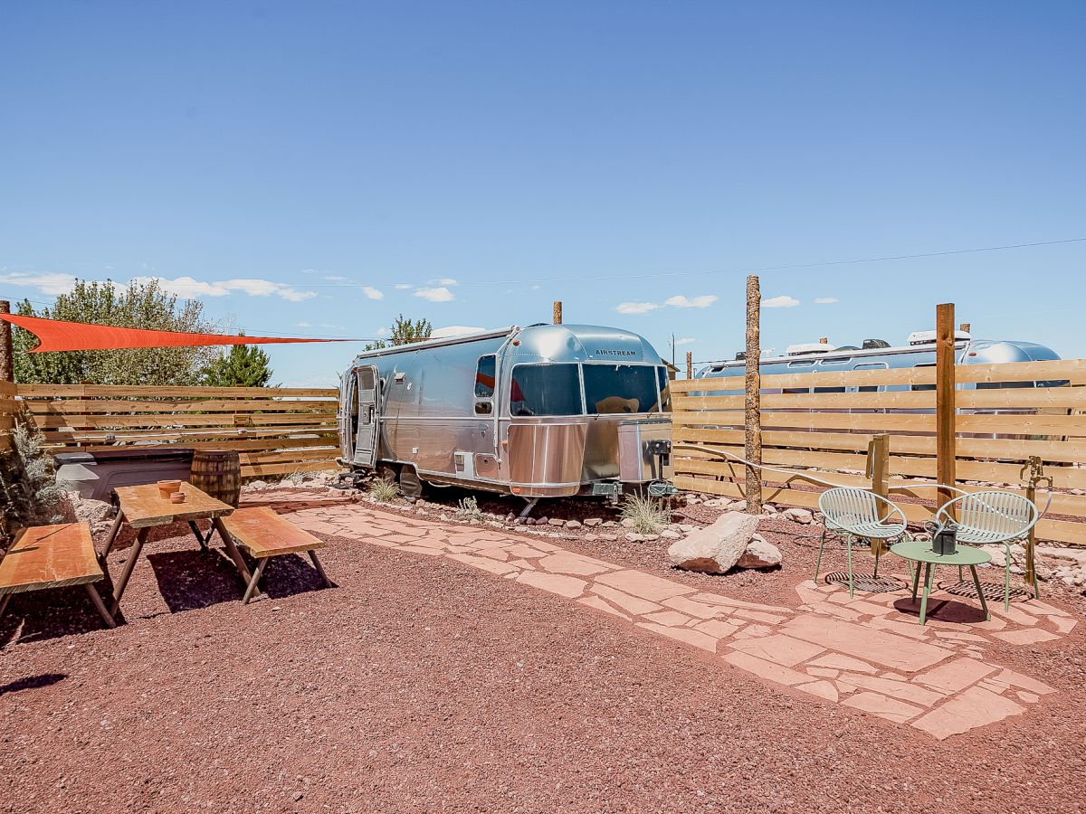 A modern Airstream trailer is parked in a fenced yard with red gravel, outdoor seating, and a picnic table under a sunny blue sky.