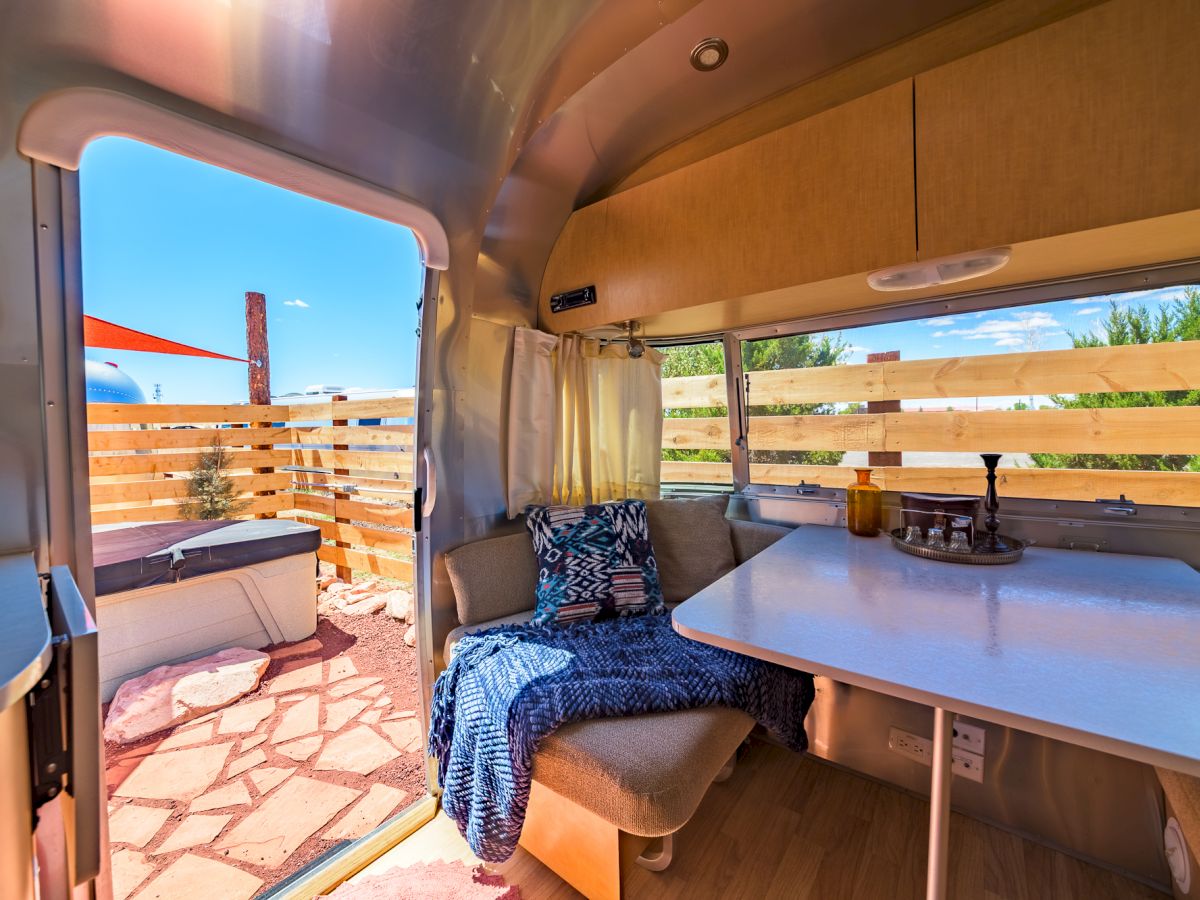 The image shows the inside of a trailer with a cozy seating area, table, and an outdoor view of a patio with wooden fence and blue sky.