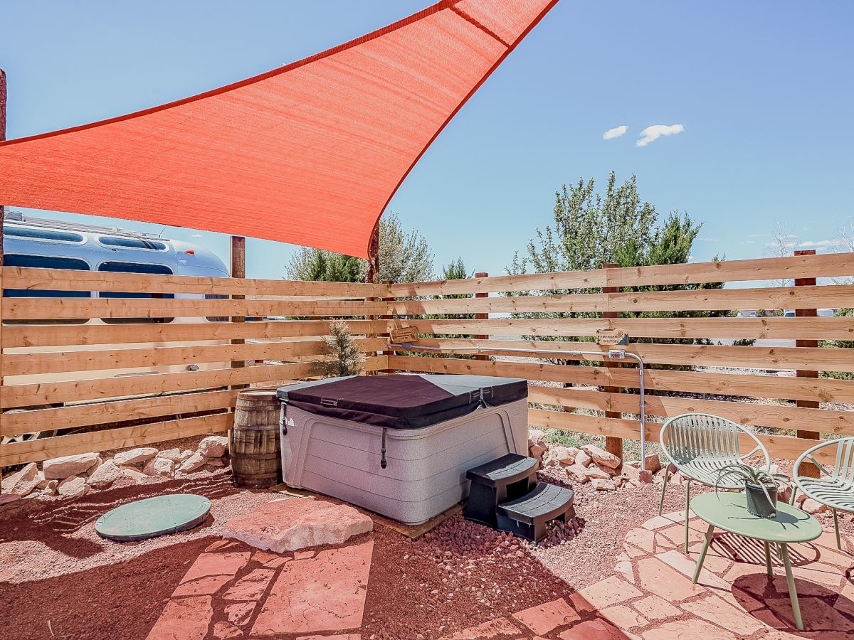 An outdoor area with a hot tub under a red canopy, surrounded by a wooden fence, chairs, table, and outdoor shower.
