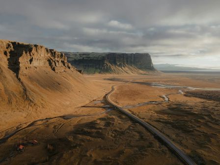 A vast landscape with a winding road and streams, flanked by rugged cliffs under a cloudy sky.