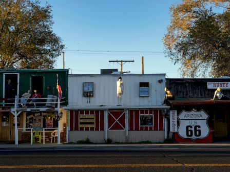 A street scene with quirky shopfronts on Route 66, mannequins, a U.S. Route 66 sign, and rustic building facades at dusk.