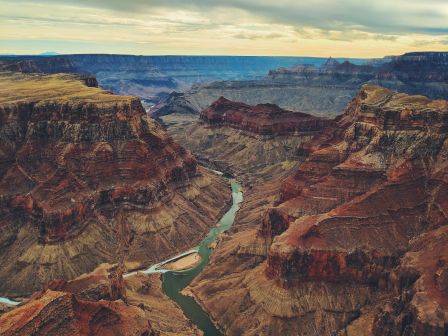A majestic view of the Grand Canyon with layered rock formations and a winding river under a cloudy sky.