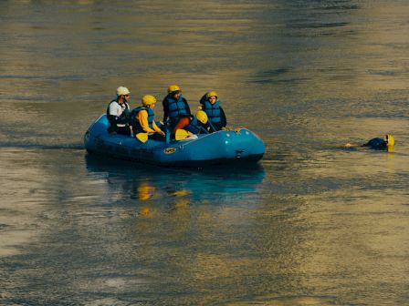 A group in life jackets is on a blue raft in the water, with one person swimming nearby wearing a helmet.