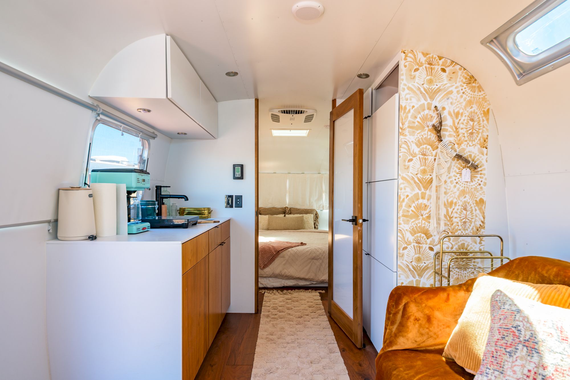 The image shows the interior of a stylish, modern camper with a kitchenette, living area, and bedroom.