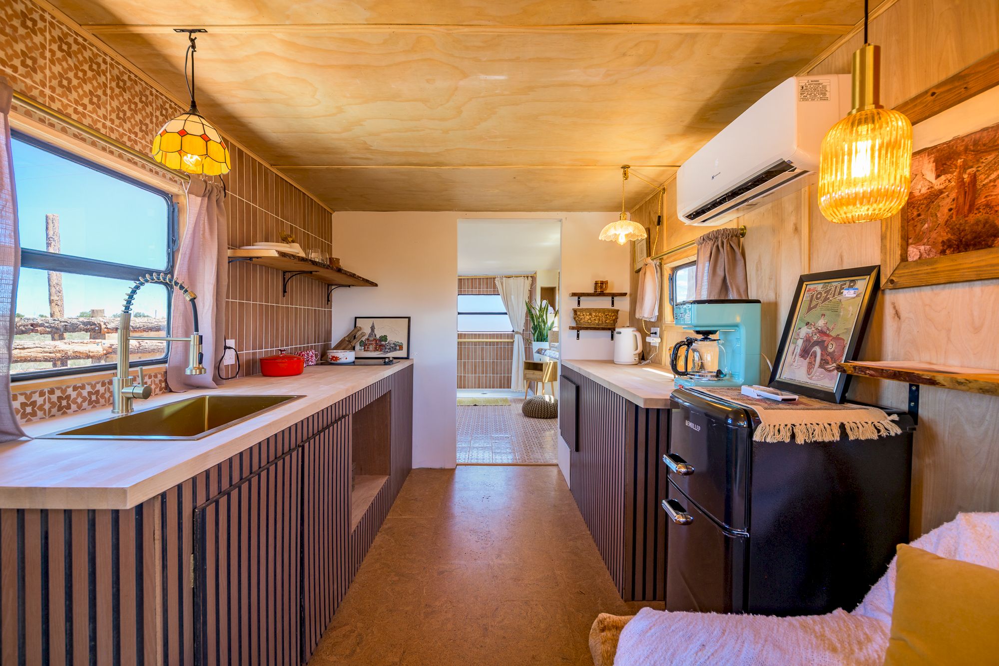 A cozy kitchen with wooden decor, modern appliances, pendant lights, and artwork on the counter; a door leads to another room.