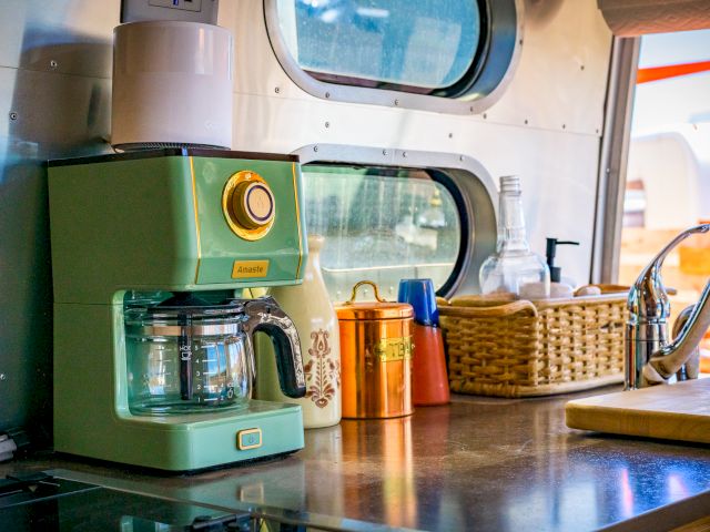 A retro green coffee maker on a kitchen counter with a mug, jars, basket, and faucet nearby in a cozy setting.