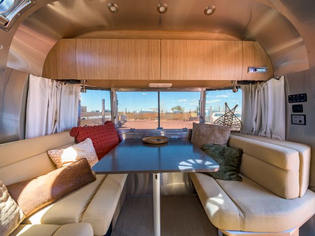 The image shows the interior of a modern camper van with a dining area, featuring beige seating, pillows, a table, and a window view.