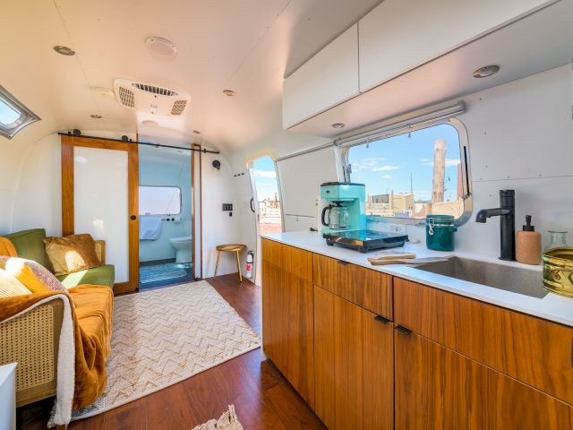 Modern RV interior with wooden cabinets, cozy seating, a rug, and kitchen appliances, bathed in sunlight from the window.