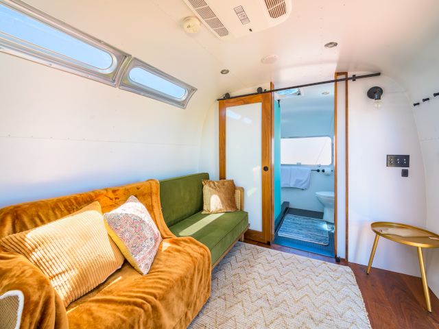 The image shows a modern tiny home interior with a green sofa, a sliding door leading to a bathroom, cozy decor, and a bright atmosphere.
