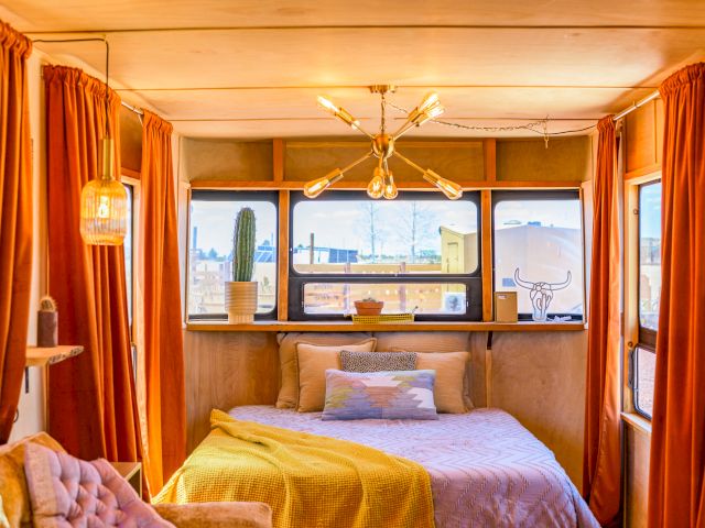 Cozy bedroom with a bed, colorful bedding, and large windows. Decor includes warm lighting, orange curtains, and cactus decor items.