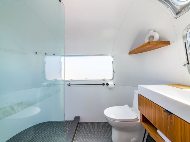 The image shows a modern bathroom in an RV with a toilet, glass partition, window, sink, and a wooden shelf with a small vase.