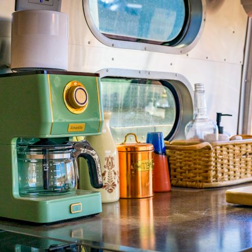 A kitchen counter with a green coffee maker, glass jar, ceramics, metal canisters, and basket under a circular window in a compact space.