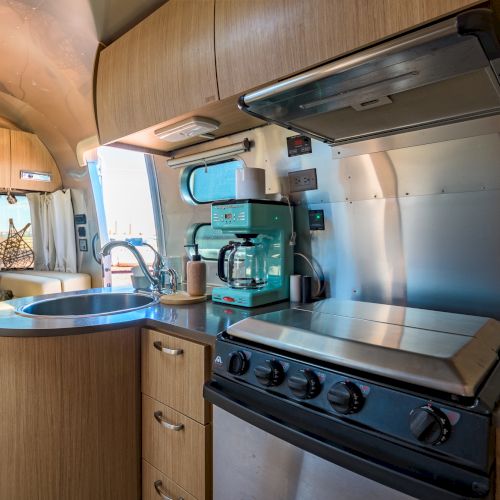 A compact kitchen inside an RV or camper with a stove, sink, and coffee maker, surrounded by wooden cabinets and a seating area.