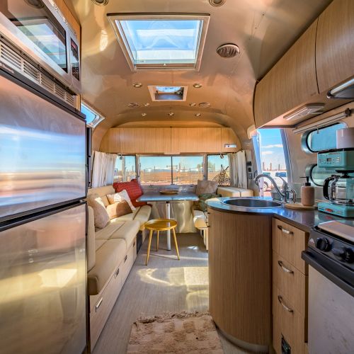 The image shows the interior of a modern camper van with a kitchenette, seating area, skylights, and large windows offering a scenic view.