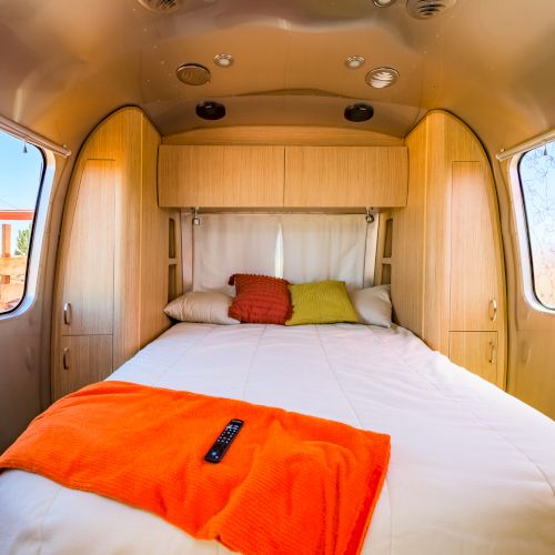 A cozy bedroom setup inside an Airstream trailer, with a bed, colorful pillows, an orange throw blanket, and a remote on top.