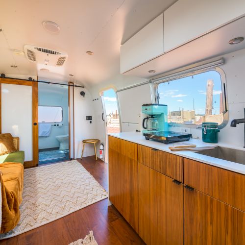 The image shows a modern camper interior with a kitchen, sofa, and bathroom, featuring wood accents and large windows.