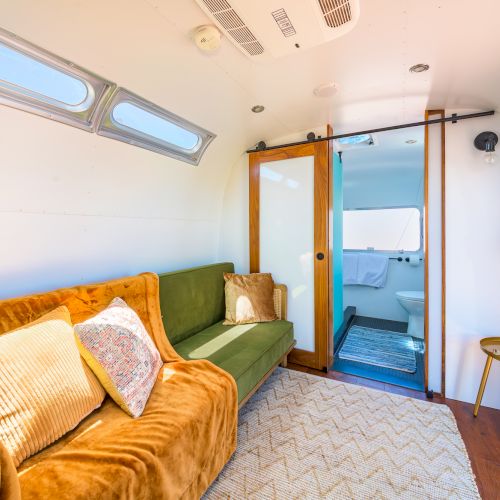 A cozy, modern tiny home interior with a green sofa, pillows, patterned rug, sliding door, and bathroom visible in the background.