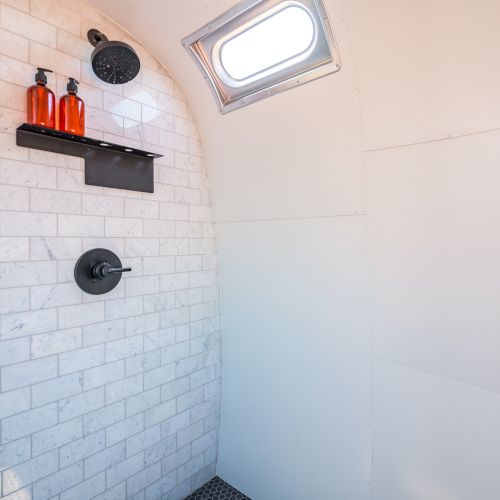 A shower with white tiles, a small window, black fixtures, and shampoo bottles on a shelf.