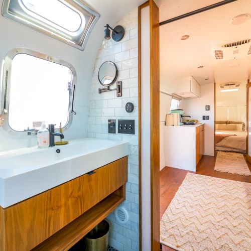A modern RV interior features a bathroom with a sink, mirror, and wood accents leading to a cozy kitchen and bedroom area.
