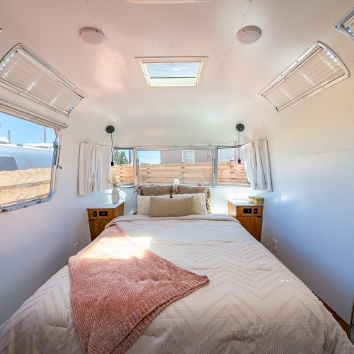 A cozy bedroom inside a trailer with a comfortable bed, wooden nightstands, a skylight, and several windows enhancing the light.