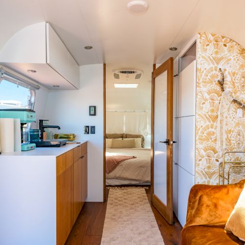 Interior of a modern, cozy trailer with a kitchenette, a brown sofa, and a bed in the background, decorated with warm tones and patterns.