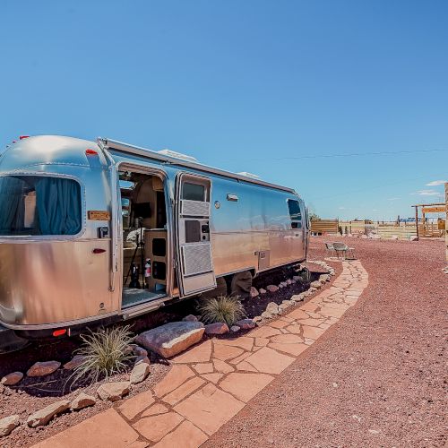 A shiny silver airstream trailer is parked on a stone pathway with a clear blue sky above.