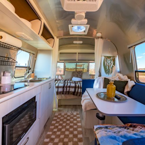 Modern RV interior with kitchen, bed, and seating area featuring a table, teal cushions, and a decorative vase. Bright and cozy setup.