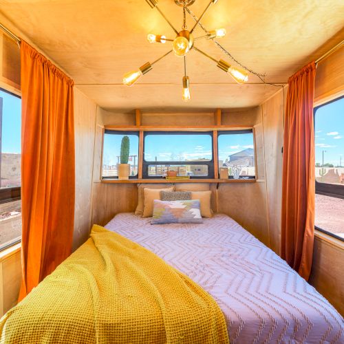 Cozy RV interior with a bed, yellow blanket, large windows, orange curtains, and a modern light fixture above.