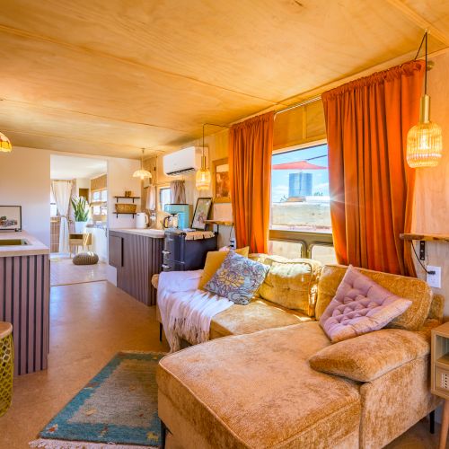 Cozy and stylish interior with a warm-toned sofa, patterned pillows, orange curtains, and kitchen area, creating a welcoming atmosphere inside.