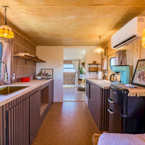 A cozy, modern kitchen with wood accents, pendant lights, a sink, and appliances, leading to a bright room in the distance.