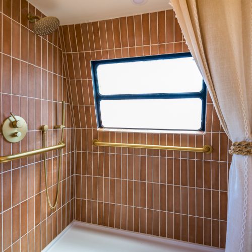 A stylish shower area with brown tiles, a window, gold fixtures, and a tied curtain.