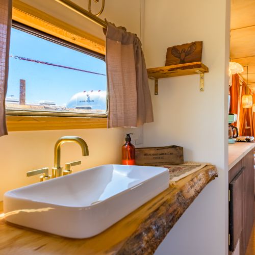 Cozy interior with a rustic sink, golden faucet, and wooden shelf. Bright, inviting living area with soft textiles and warm lighting at the end.
