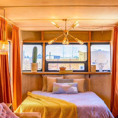 A cozy room with orange curtains, a bed with colorful cushions, a cactus on the windowsill, and a unique chandelier lights the space.