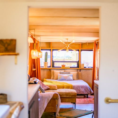 A cozy interior view featuring a warmly lit bedroom with a bed, sofa, and decorative lighting, seen through a doorway with gentle natural light.