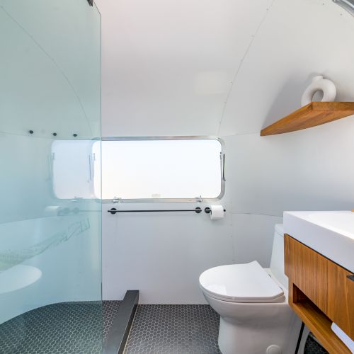 The image shows a modern bathroom with a toilet, sink, wooden cabinet, and large window. There's also a glass shower partition.