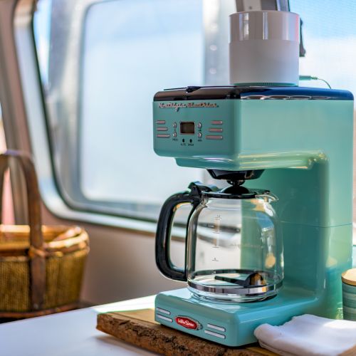 The image shows a retro-style turquoise coffee maker on a countertop with a glass carafe, next to a basket and a window.