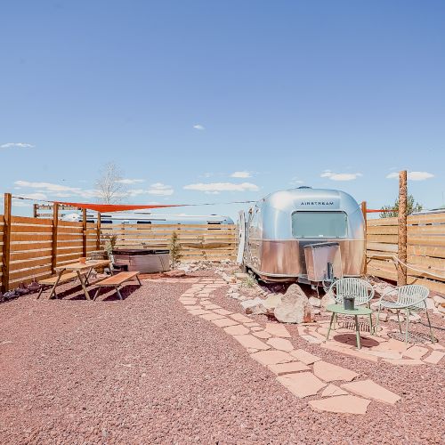 The image shows an Airstream trailer in a fenced yard with pink gravel, patio furniture, a picnic table, and a path under a clear blue sky.