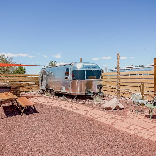 A vintage Airstream trailer is parked in a fenced area with outdoor seating and a wooden table on a sunny day.