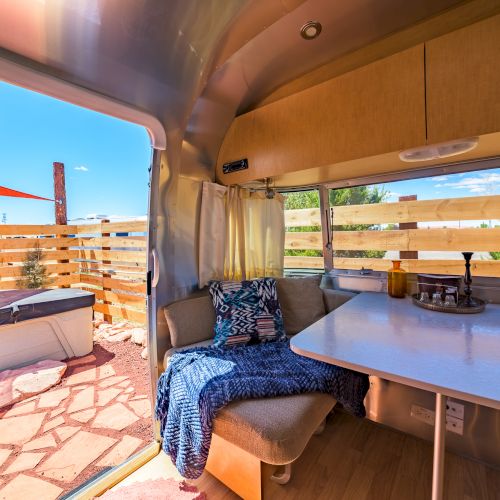 Interior view of a cozy trailer with a table, seating, and open door showing an outdoor patio with a hot tub surrounded by fencing.