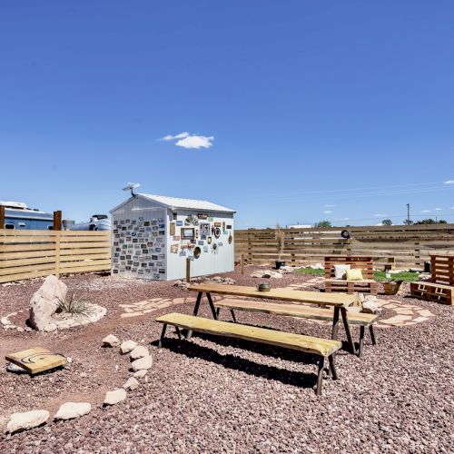 The image shows a fenced outdoor area with picnic tables, a small shed, and gravel ground, under a clear blue sky.