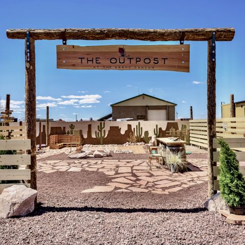 The image shows a rustic wooden gate labeled "The Outpost," leading to a desert-themed patio area with cacti and a central structure.