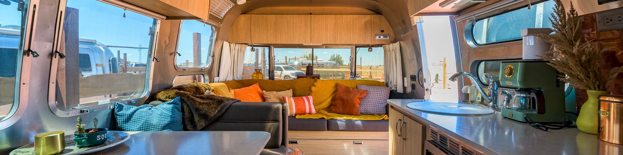 The image shows the interior of a cozy, well-lit camper van with a kitchen, seating area, and decorative cushions.