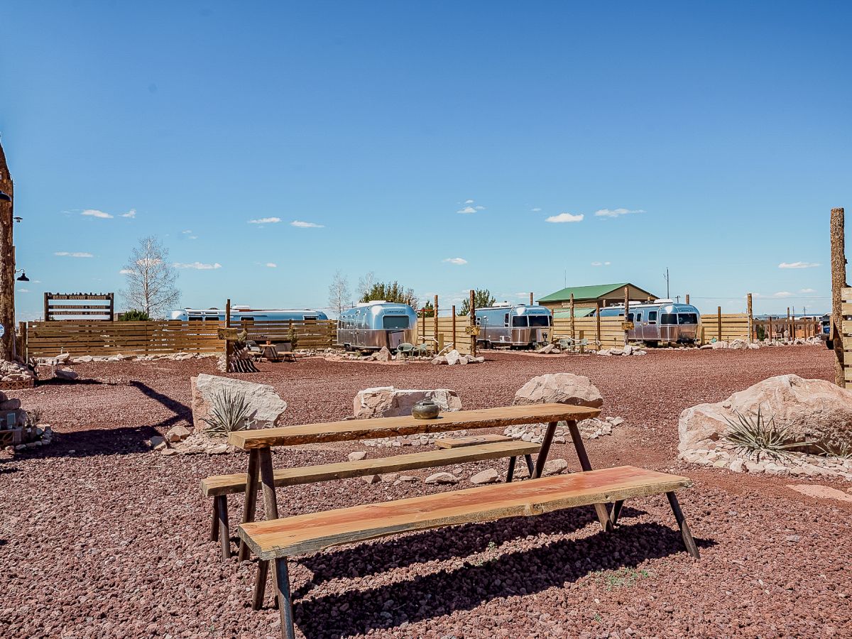 The image shows a rustic outdoor area with picnic tables, wooden fences, rocks, and trailers under a clear blue sky.