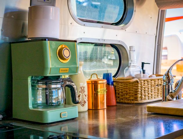 A retro green coffee maker on a kitchen counter with a mug, jars, basket, and faucet nearby in a cozy setting.