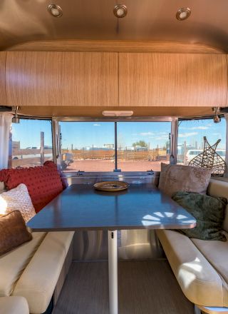 The image shows the interior of a modern camper van with a dining area, featuring beige seating, pillows, a table, and a window view.